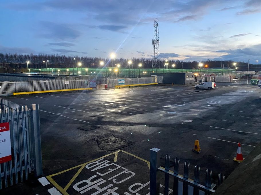 Empty industrial area at dusk, illuminated by floodlights, with parked vehicles and a tower.