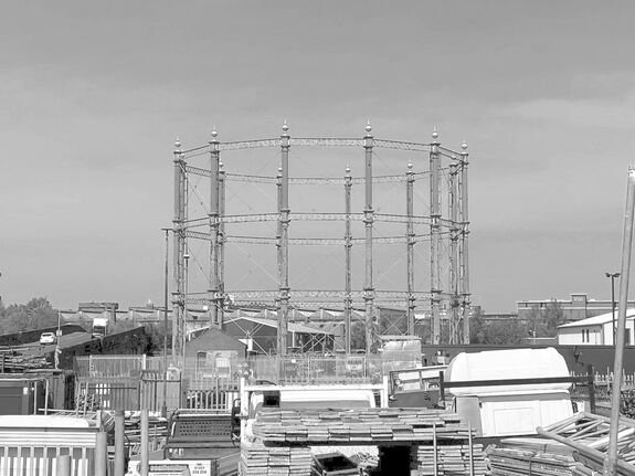 Gasometer structure surrounded by industrial buildings under a clear sky.