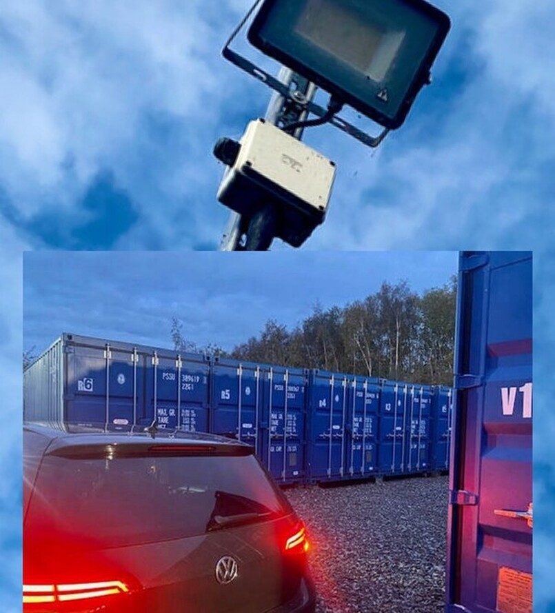 Spotlight illuminating a vehicle near blue shipping containers at dusk.