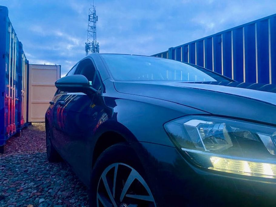 A dark car parked between shipping containers under a cloudy sky.