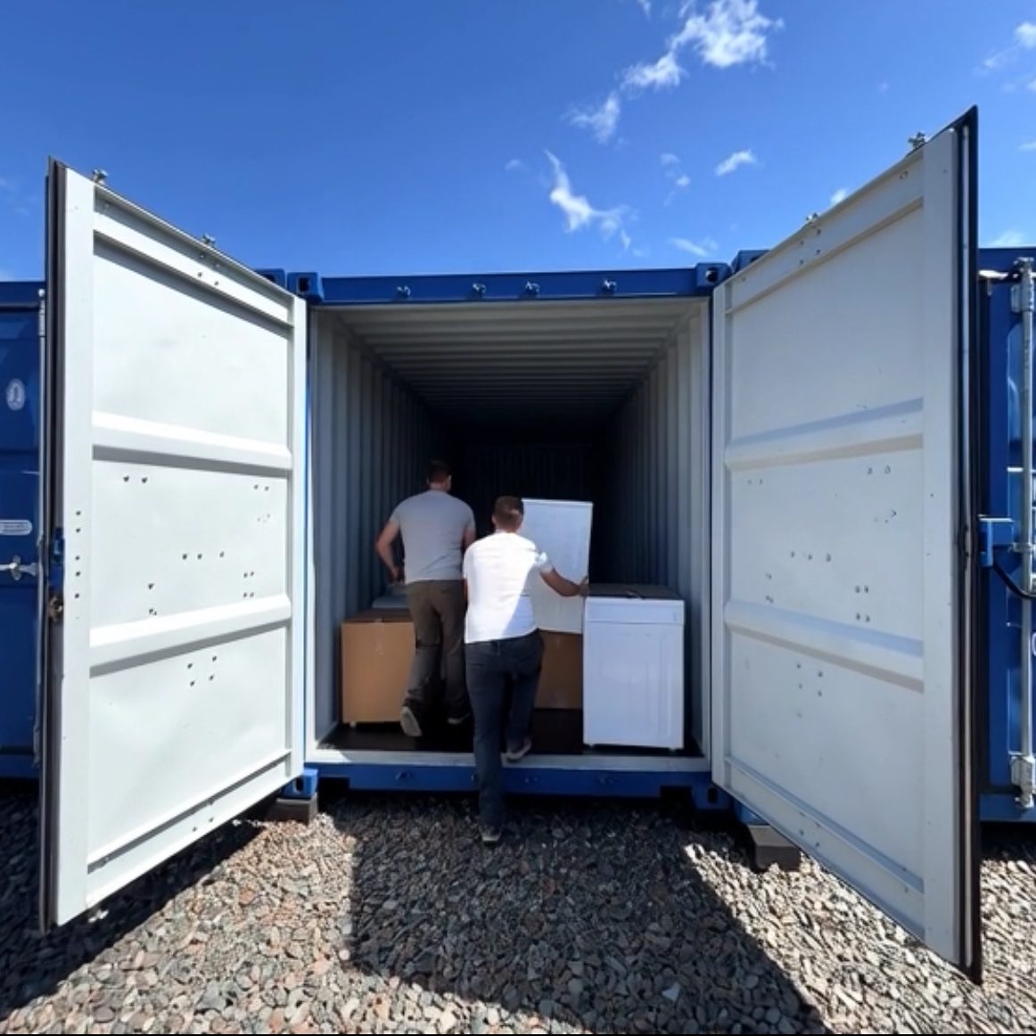 Two people loading boxes into a blue shipping container under a clear sky.