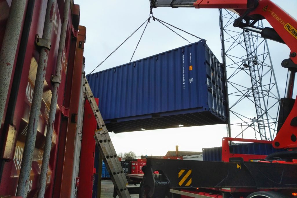A blue shipping container being lifted by a crane at a loading dock.