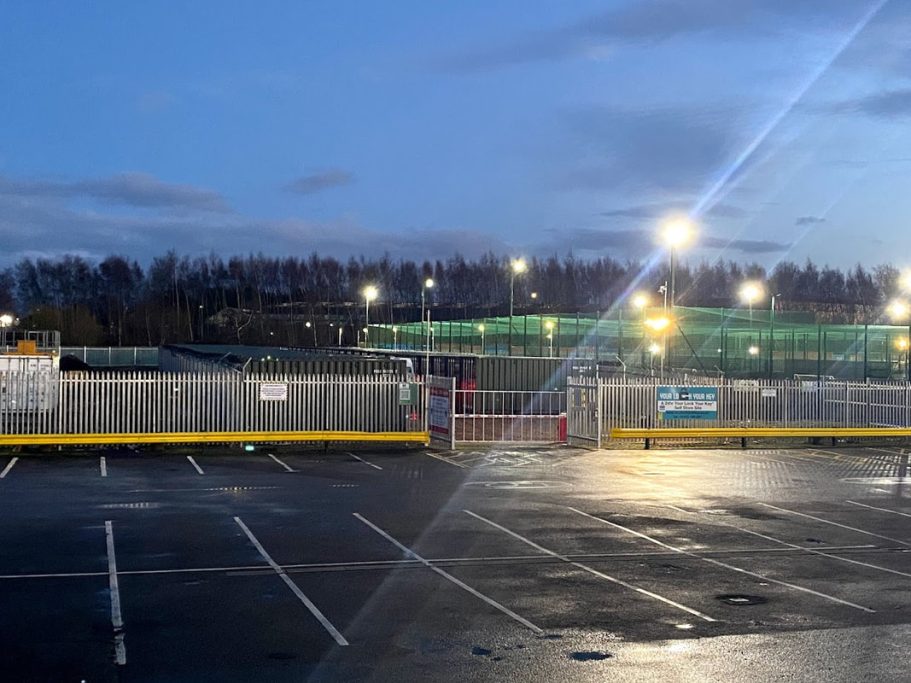Empty car park at dusk, with illuminated fencing and construction in the background.