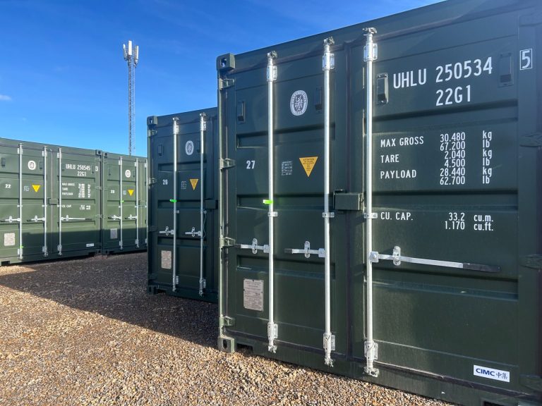 Row of green shipping containers on a gravel surface under a clear blue sky.