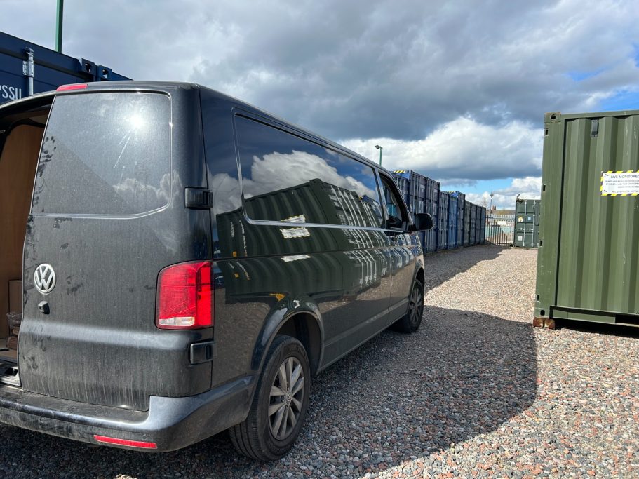 Black van parked in a gravel area surrounded by shipping containers under a cloudy sky.