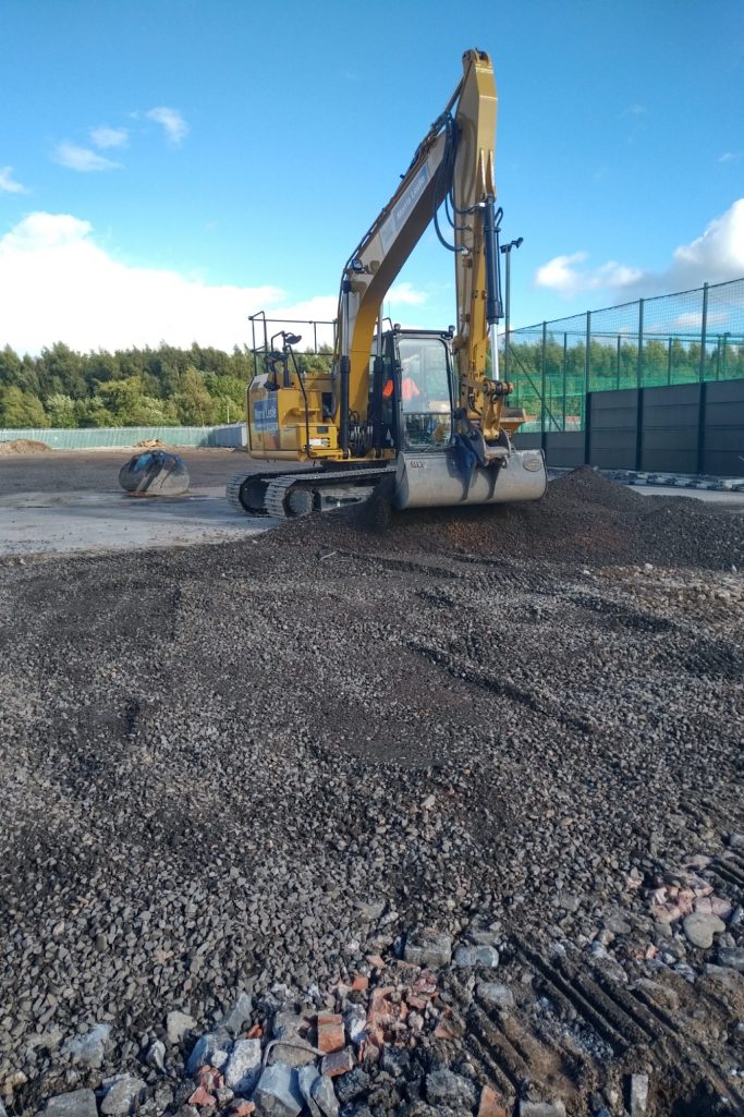 A yellow excavator operates on a construction site with gravel and a fence in the background.