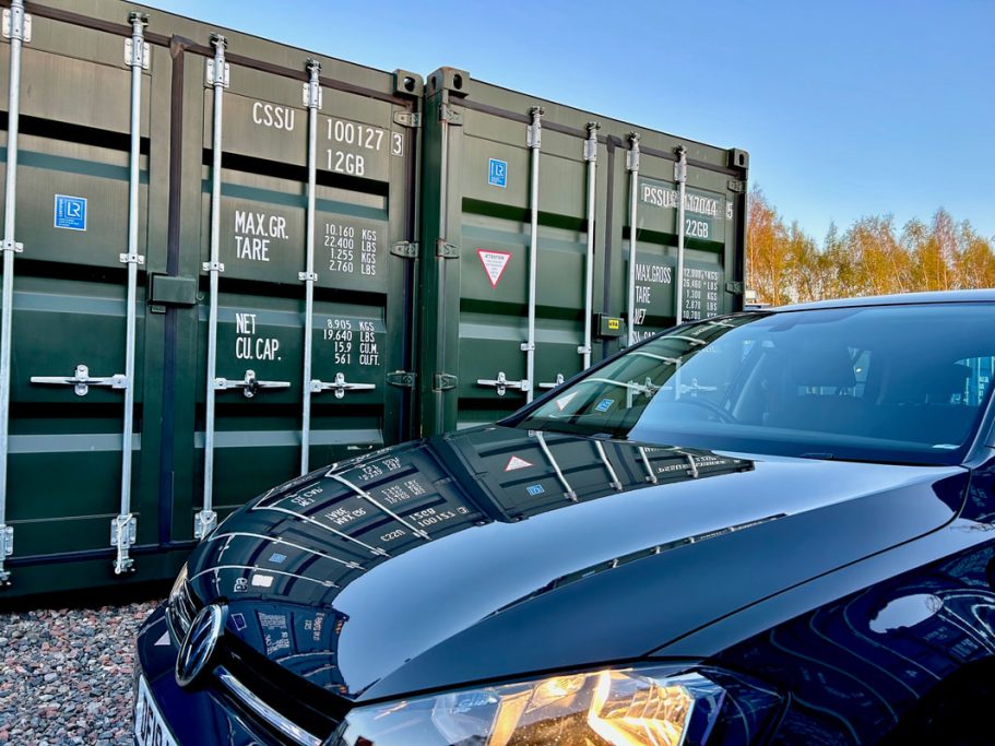 Black car parked in front of green shipping containers under a clear blue sky.
